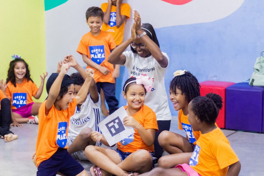 um grupo alegre de crianças e uma mulher em um ambiente interno, uma sala de aula ou espaço educativo. As crianças estão sentadas no chão, sorrindo, rindo e comemorando com entusiasmo. Muitas delas vestem camisetas laranja com a logo 'Cores do Mará', enquanto a mulher, que também sorri e bate palmas com os braços erguidos, usa uma camiseta branca com a frase: 'Eu transformo vidas no Maranhão'