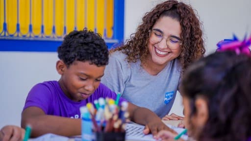 Educadora sorrindo olhando para aluno empolgado fazendo uma atividade escrita sobre a mesa.