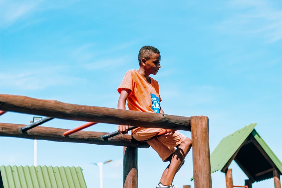 A black boy playing on a wooden playground outdoors, under a clear and sunny blue sky. He is sitting on a log structure, with his legs on one side and his torso on the other, in a pose that suggests movement and fun. The boy is wearing an orange t-shirt and shorts of the same color, with sports sneakers. On the t-shirt, there is the institute's logo in blue, in the shape of a São João flag, containing the text 'CORES DO MARÁ' and a star. In the background, green roofs of other playground structures are visible. The image has rounded corners and colorful little flags in the corners — reinforcing a cheerful and childlike tone.