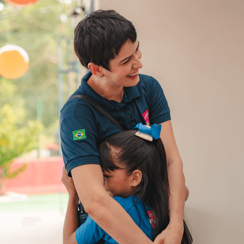 Mulher sorrindo abraçando uma menina em ambiente escolar ou recreativo, transmitindo acolhimento, carinho e cuidado.